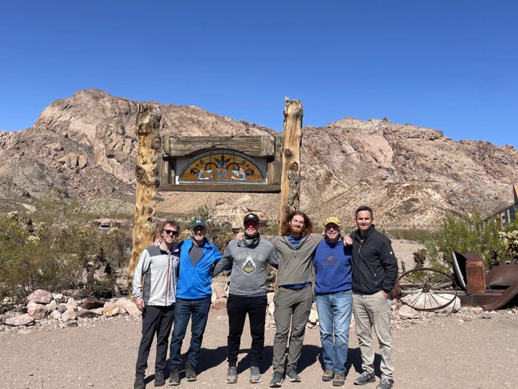 Six friends pose arm in arm by a wooden sign, smiling with the stunning Nevada desert and Colorado River near Las Vegas as backdrop.