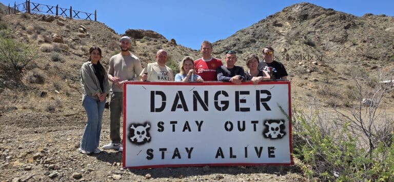 Eight people near Las Vegas pose by a DANGER STAY OUT sign in the Nevada desert—ideal for RZR rides & ghost town tours.