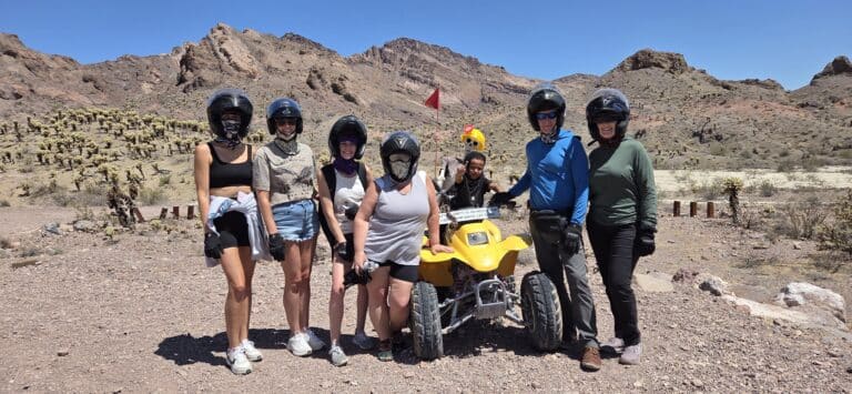 Seven adventurers in helmets pose by a yellow ATV in Nevada’s desert near Las Vegas during an epic ATV tour.
