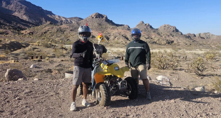 Two riders in helmets pose by a yellow ATV near Nevada’s rocky desert, ready for Las Vegas RZR off-road and Ghost Town tours.