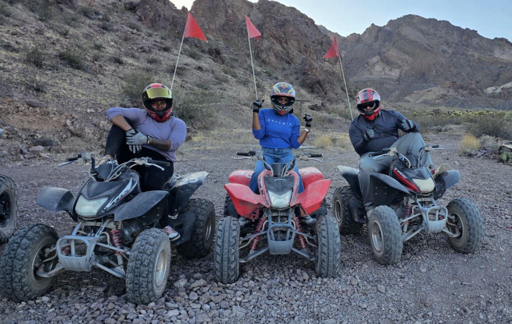 Three riders in helmets and gloves pose on ATVs with red flags, ready for an off-road Las Vegas desert or Nevada ATV tour.
