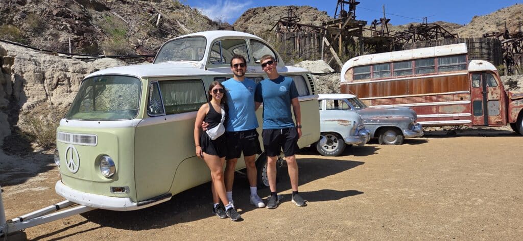 Three friends smile by a vintage VW van in a Nevada ghost town near Las Vegas, with classic cars and desert ATV adventures nearby.