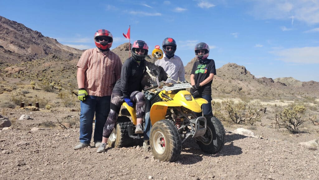 Group in helmets poses with a yellow ATV in Nevada’s rocky desert near Las Vegas—perfect for ATV tours or RZR off-road adventures.