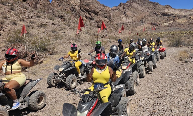 Adventurers in yellow gear ride ATVs on a Nevada desert trail near Las Vegas with red flags, mountains, and dry bushes.