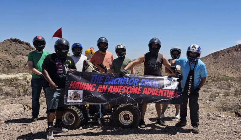 Eight helmeted friends in Nevadas desert hold a ‘Bachelor Crew Adventure’ banner by a red ATV on a Las Vegas RZR tour.