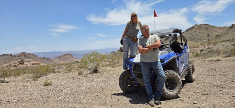 A man stands by a blue RZR in Nevada desert near the Colorado River, woman on hood—perfect for Las Vegas ATV tours and Ghost Town sightseeing.