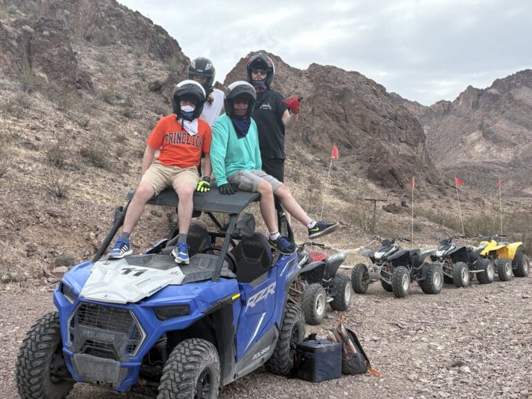 Adventurers in helmets enjoy an ATV tour near the Colorado River in Nevada’s rocky desert outside Las Vegas, with RZRs and mountains behind.