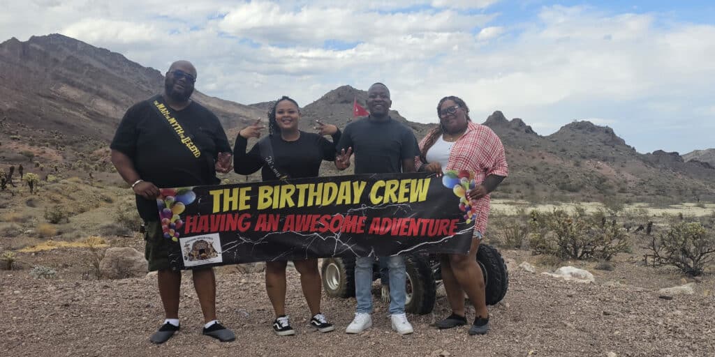 Four friends pose with a Birthday Crew banner in the Nevada desert near Las Vegas after an epic RZR off-road adventure.