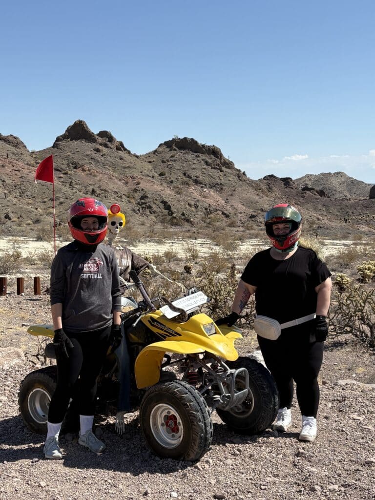 Two riders in helmets with a skeleton sit on a yellow ATV in the Nevada desert near Las Vegas; perfect for ATV tours & ghost town adventures.