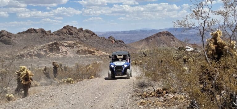 A blue RZR off-road vehicle cruises a rocky desert trail near Las Vegas, Nevada, with cacti and rugged hills in the backdrop.