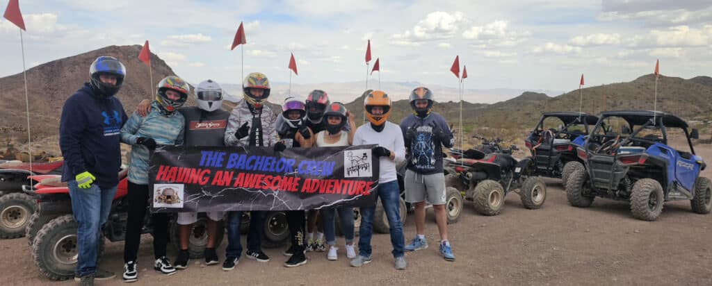 A group near Las Vegas holds a “Bachelor Crew Adventure” banner before ATVs, ready for RZR rides and Ghost Town tours by the Colorado River.