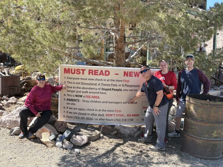 Four friends smile by a rules sign at a Nevada ghost town near the Colorado River—perfect for RZR rides and ATV tours.