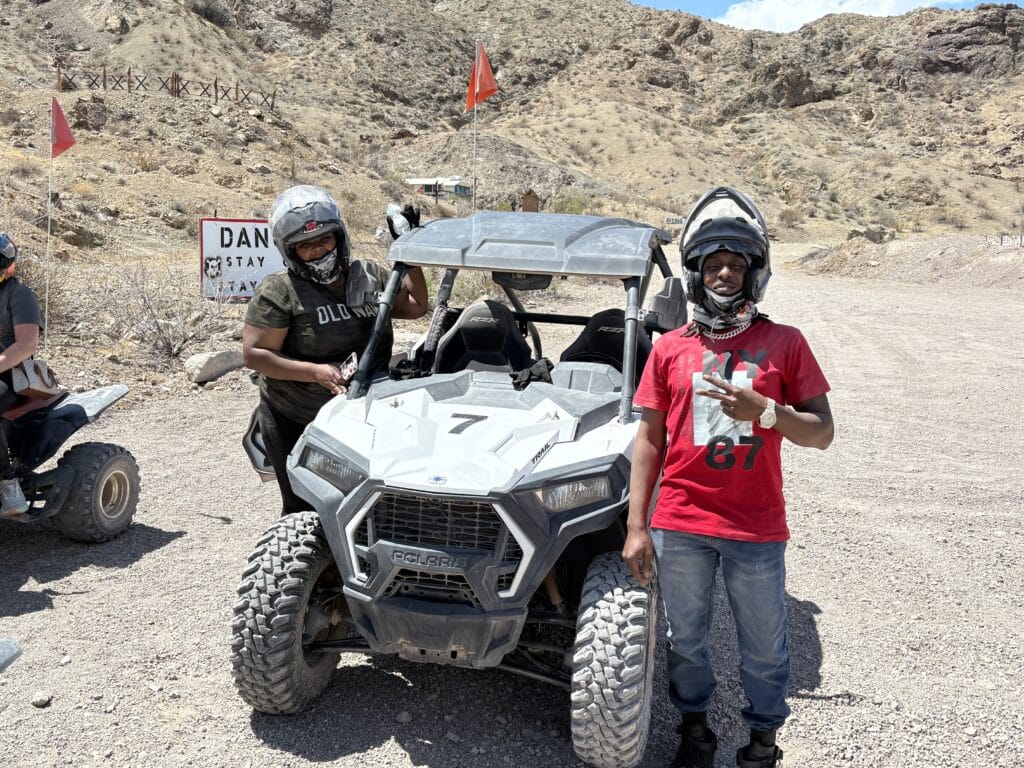 Adventurers in helmets beside a white RZR on a rocky Nevada trail near Las Vegas, with a Danger sign and desert hills in view.