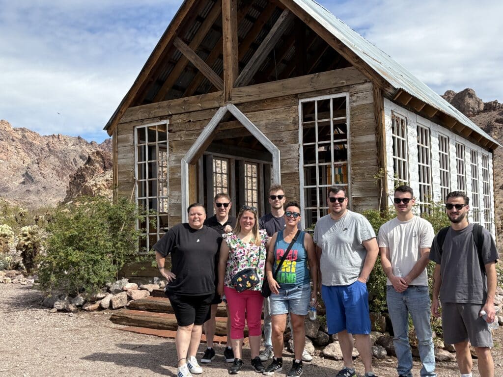 Seven friends smile in front of a rustic wooden building near Las Vegas on a Ghost Town sightseeing and RZR off-road adventure.