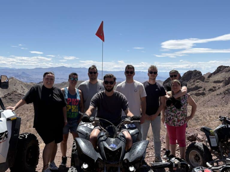 A group enjoys an ATV tour near Las Vegas, Nevada, posing on rocky terrain by the Colorado River with mountains and an orange flag.