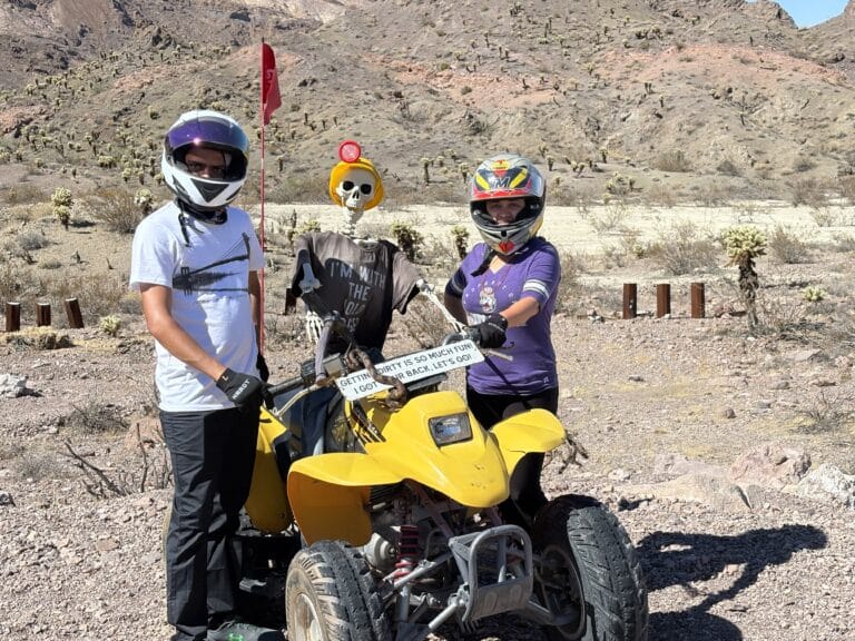 Two riders in helmets pose by a yellow ATV near Las Vegas with a quirky skeleton at a Nevada desert ghost town ATV tour.