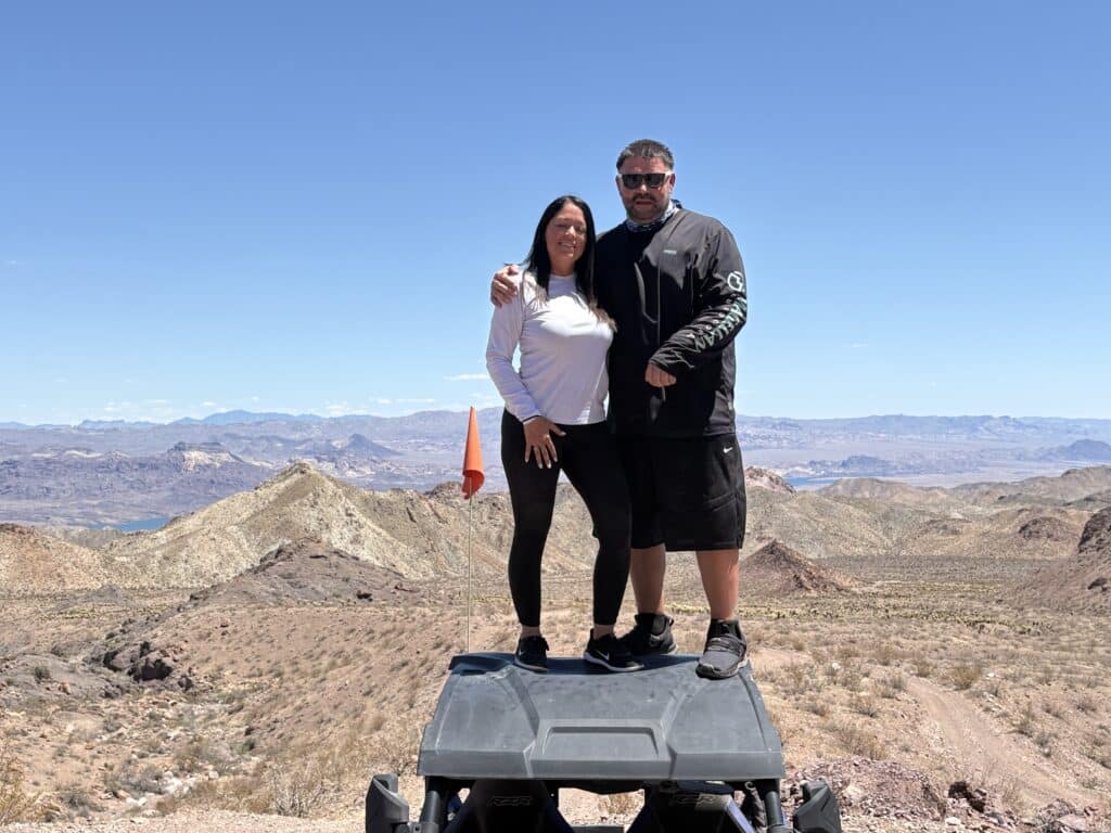 Smiling couple atop an RZR in Nevadas desert near Las Vegas, with Rocky mountains—perfect for ATV tours & Ghost Town sightseeing.