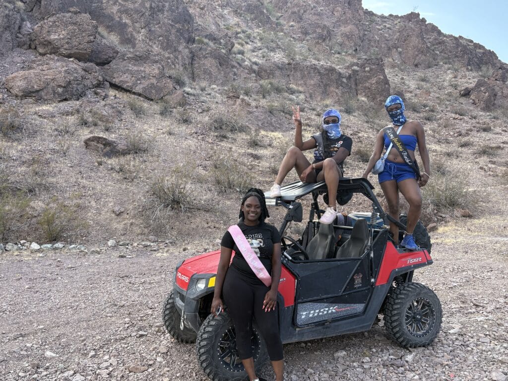 Three women in sashes pose by a red RZR on Nevada desert rocks near Las Vegas, ready for an ATV tour along the Colorado River.