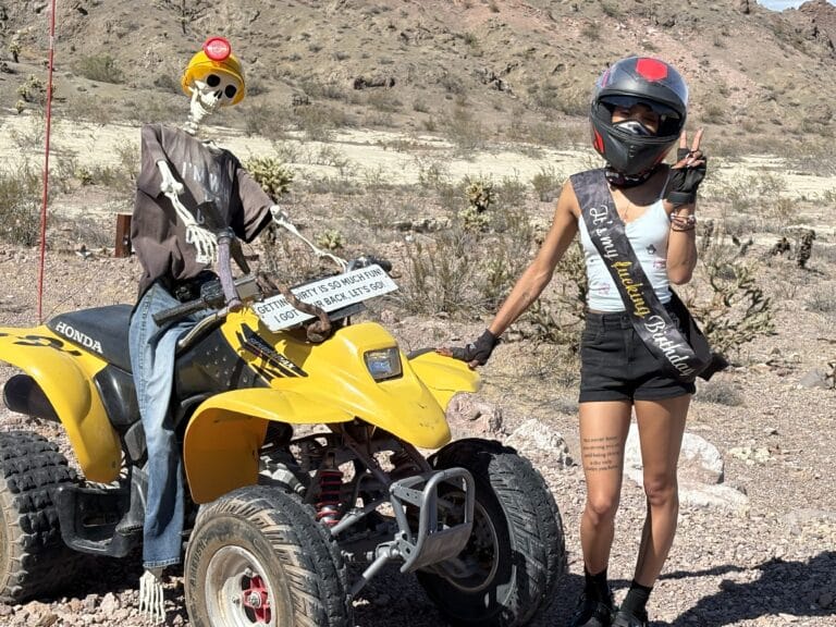 Woman in helmet poses with peace sign by yellow ATV and skeleton in Nevada desert near Las Vegas Ghost Town ATV tour.