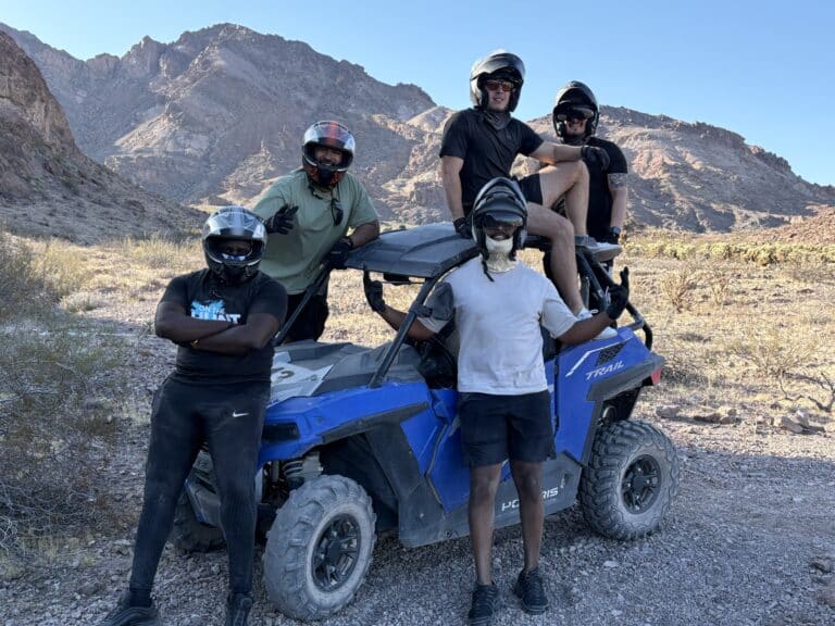 Five friends in helmets pose on a blue RZR during an ATV tour near Las Vegas, Nevada, with desert mountains in the background.