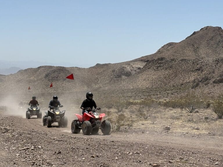 Adventure seekers enjoy an ATV tour near Las Vegas, riding single file on a dusty Nevada desert trail with red flags flying.