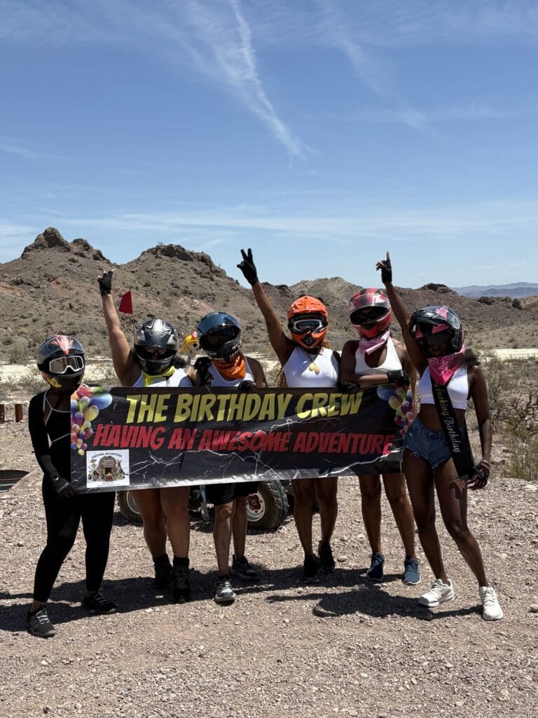 Six adventurers in helmets pose with ‘The Birthday Crew’ banner during a Nevada ATV tour near the Colorado River by Las Vegas.