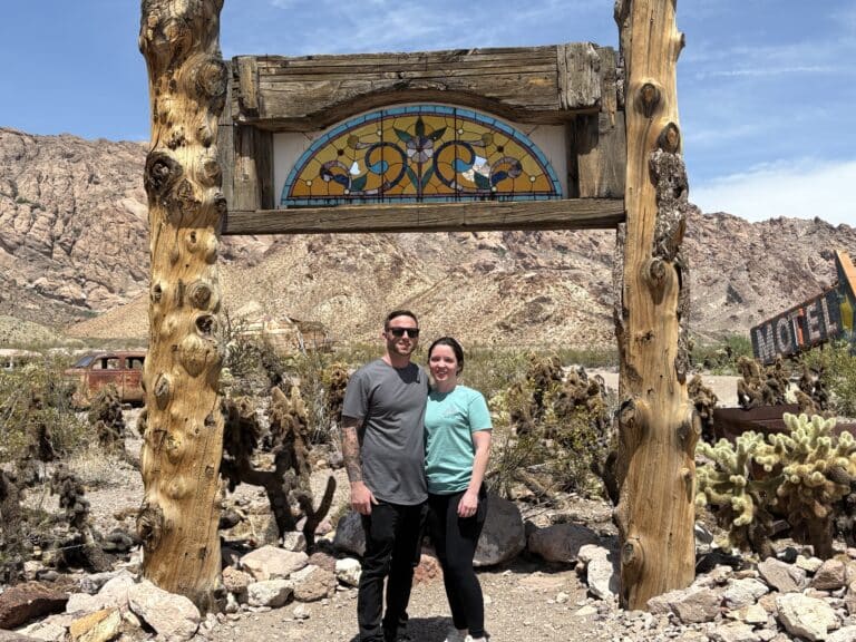 Smiling couple under rustic sign near Las Vegas, Nevada, with desert views—perfect for Ghost Town tours & ATV adventures.
