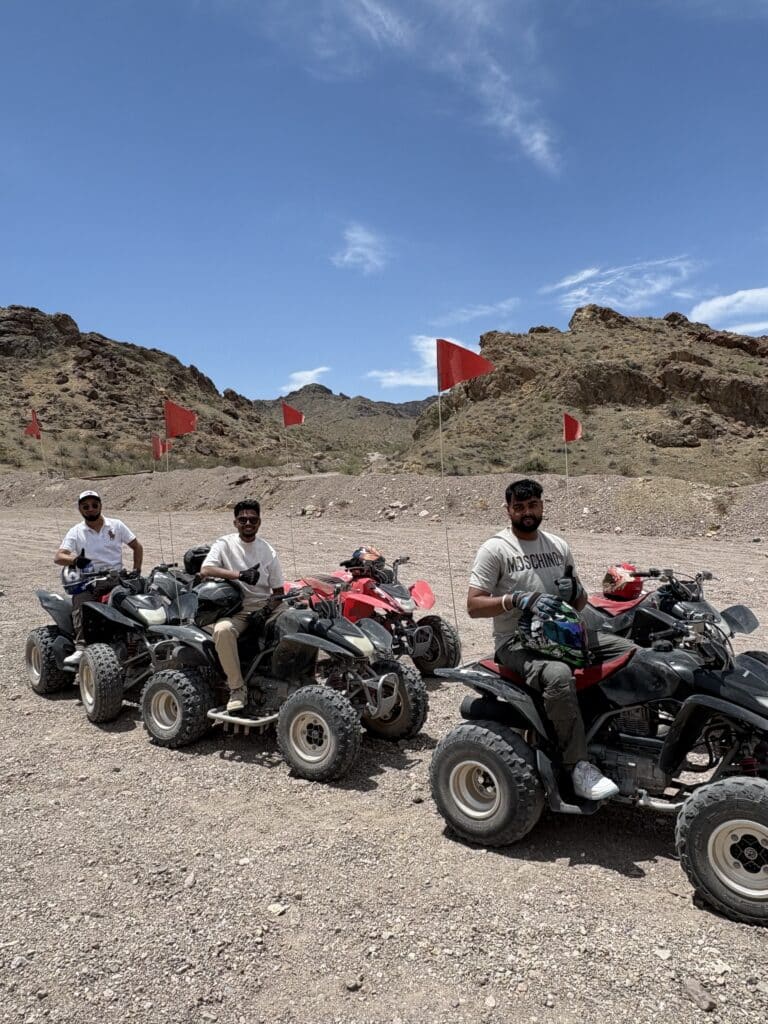 Three men in sunglasses enjoy an ATV tour near Las Vegas, Nevada, riding quad bikes in the desert with red flags waving.