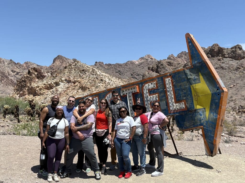 A group of eleven poses by a vintage motel sign near Las Vegas, Nevada, with desert mountains—perfect for ATV tours and Ghost Town sightseeing.