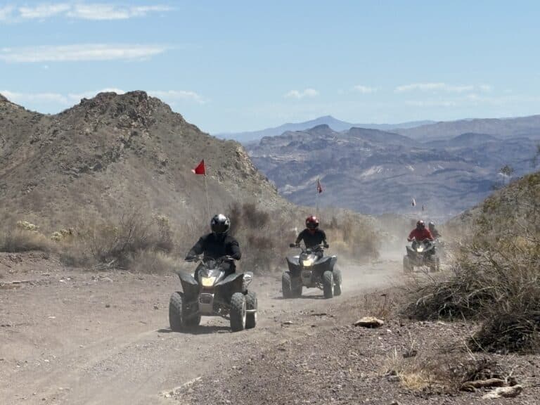 Three riders on ATVs explore a dusty Nevada desert trail near Las Vegas, with red flags waving and mountains in the background.