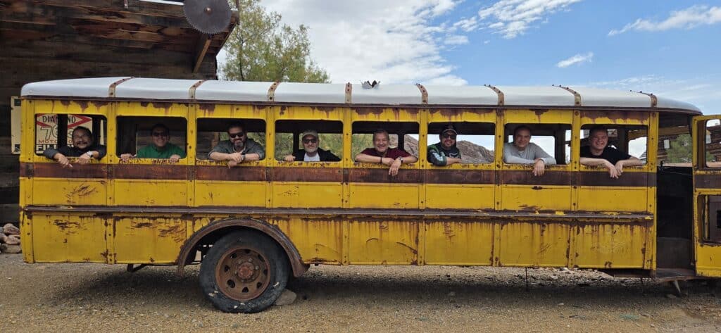 Eight friends smile from a rusty, wheel-less school bus in a Nevada ghost town near the Colorado River—perfect for ATV tours!.