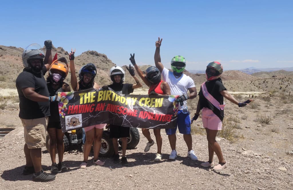 A group in helmets and casual clothes celebrates with a Birthday Crew banner during an ATV tour near the Colorado River, Nevada.