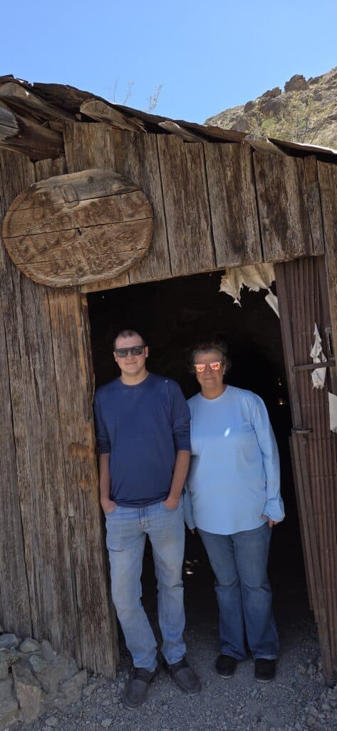 Two friends in sunglasses pose by a rustic Nevada ghost town cabin near the Colorado River—perfect for Las Vegas ATV tours!.