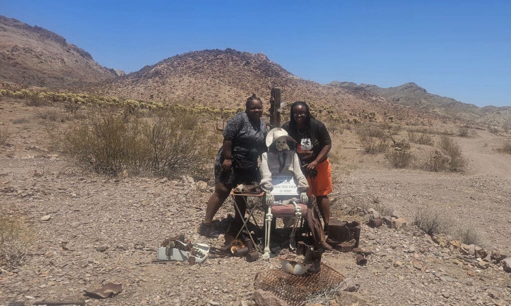 Smiling friends pose by a skeleton in a hat and shades during a Nevada ghost town ATV tour near the Colorado River, Las Vegas.