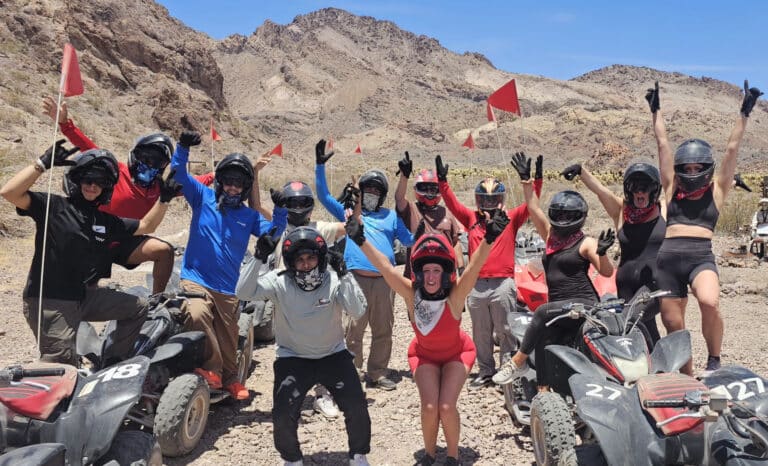 Excited group on a Las Vegas ATV tour poses with RZRs and red flags in Nevada’s rocky desert near the Colorado River.