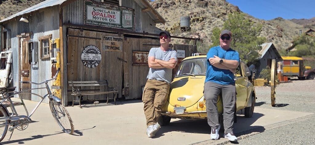 Two men lean on a vintage yellow car by a rustic Nevada garage near Las Vegas, perfect for Ghost Town sightseeing and ATV tours.