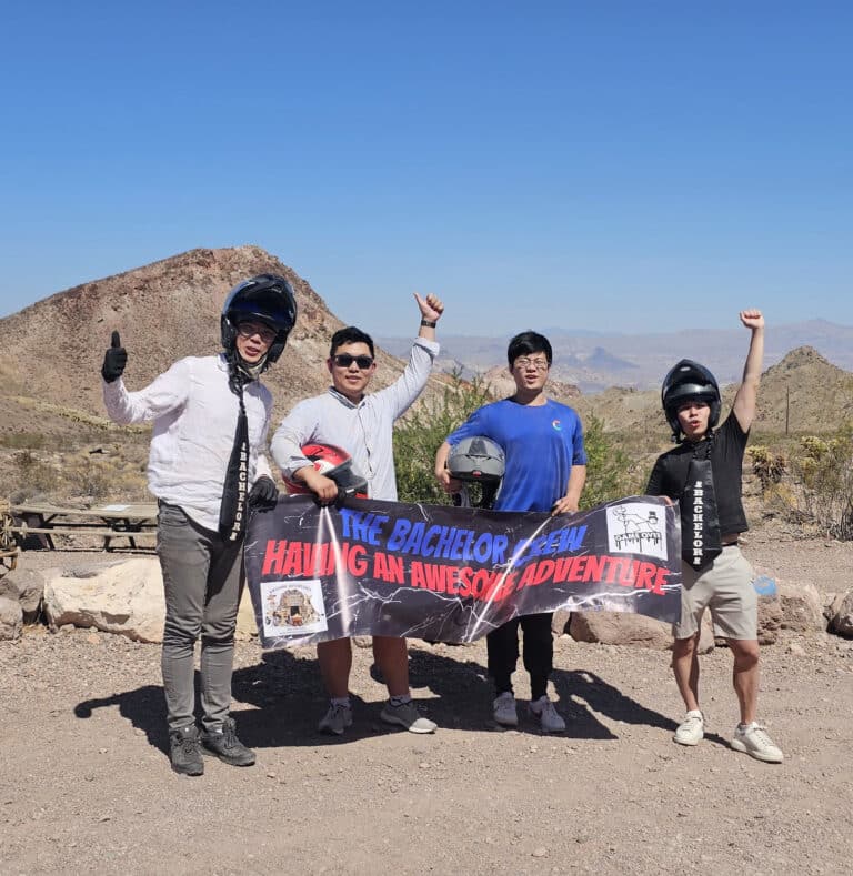 Four men in helmets pose with a Bachelor Team Having an Awesome Adventure banner on a Las Vegas ATV tour near the Colorado River.