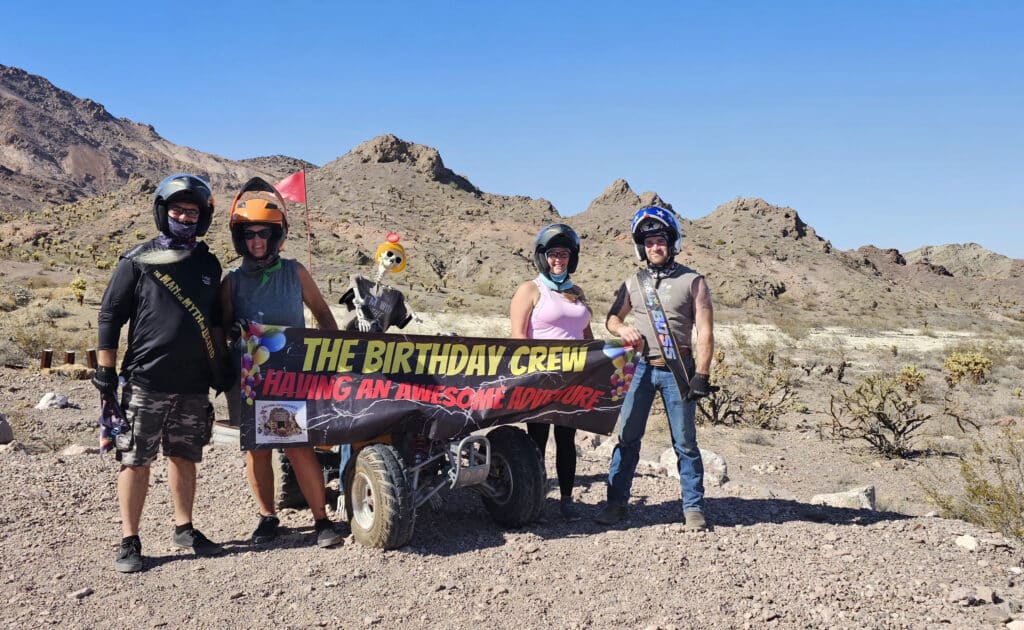 Four friends in helmets pose by an ATV, holding a Birthday Crew banner on a Las Vegas RZR tour near the Colorado River.