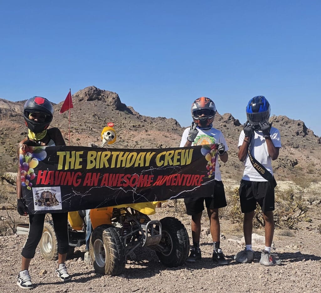 Three friends in helmets pose with a banner during an ATV tour near Las Vegas, Nevada, with mountains and a yellow RZR behind them.