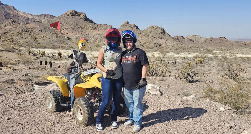 Smiling duo in helmets pose by a yellow ATV near Las Vegas, Nevada—perfect for Colorado River ATV tours and Ghost Town rides.