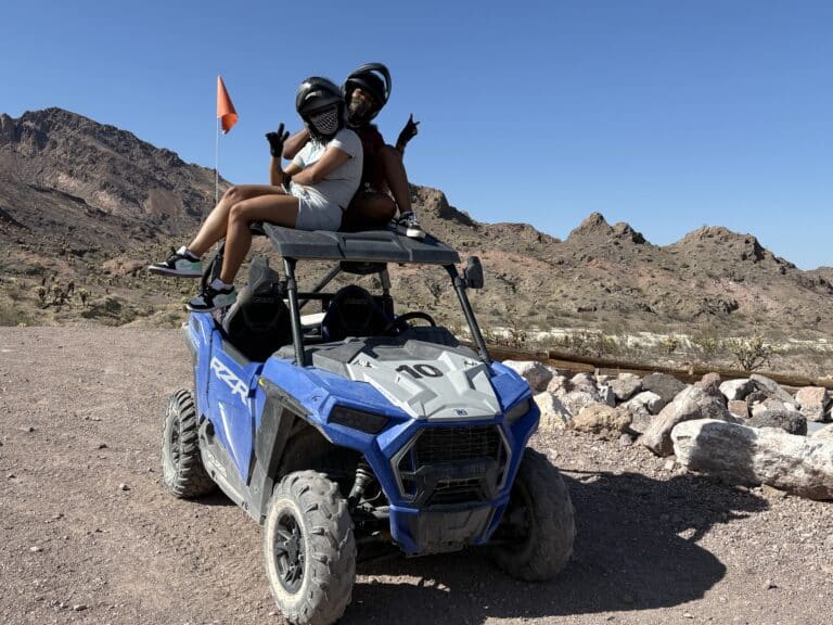 Adventurers in helmets pose with peace signs atop a blue ATV in Nevada’s desert near Las Vegas on an exciting RZR off-road tour.