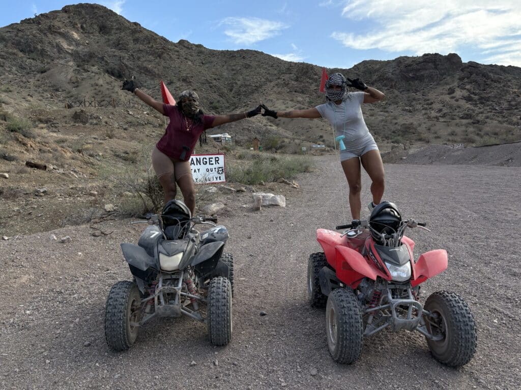 Two adventurers pose on ATVs in the Nevada desert near Las Vegas, enjoying an ATV tour with rocky hills and a DANGER sign behind them.