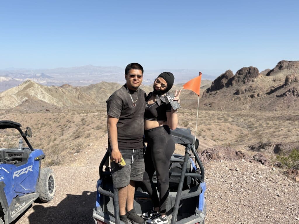 Smiling duo pose atop a blue RZR in Nevada’s desert near Las Vegas, with mountains behind—perfect for ATV tours and Ghost Town sights!.
