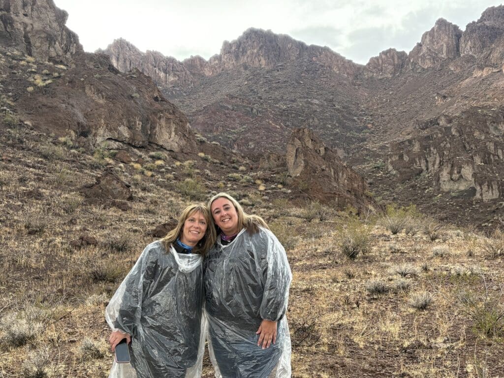 Two women in gray rain ponchos smile in Nevada’s rocky desert, enjoying a Las Vegas ATV tour near the Colorado River.