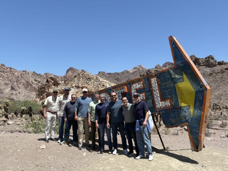 Ten men smile in front of a vintage arrow sign in Nevada’s rocky desert near Las Vegas, ready for RZR off-road and Ghost Town tours.