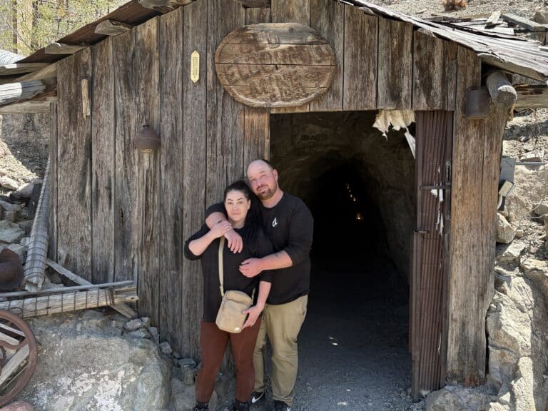 Smiling couple at Nevada mine entrance near Colorado River—surrounded by rocks, relics & old tools. Perfect for Las Vegas ATV tours!.