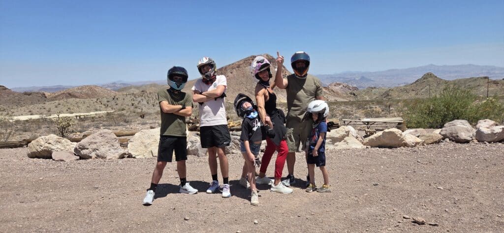 Group in helmets and casual wear poses on Nevada ATV tour path near Las Vegas, mountains, and Colorado River backdrop.