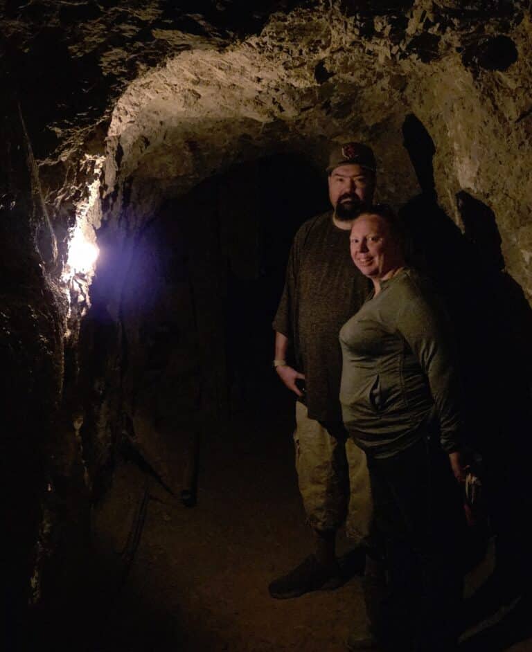 Two explore a dim, rocky tunnel near the Colorado River in Nevada, shadows cast by their ATV tour light in Las Vegas.
