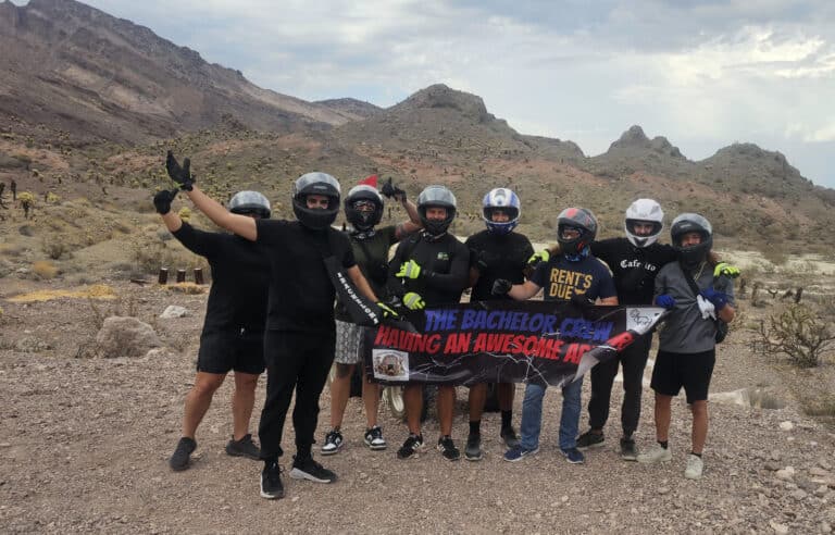 Nine friends in helmets pose with ‘THE BACHELOR CREW HAVING AN AWESOME ADVENTURE’ banner on a Las Vegas Nevada ATV tour.