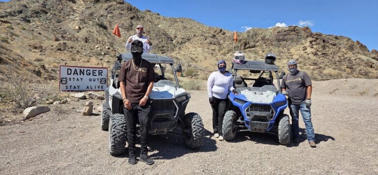 Group in bandanas and sunglasses by ATVs near Las Vegas, Nevada, pose on rocky terrain with Stay Out sign; RZR off-road adventure.